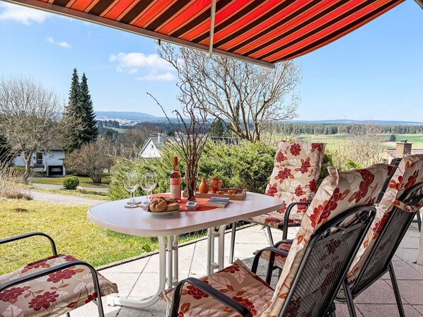 En udendørs patio med et bord dækket til to, omgivet af blomstrede stole, med udsigt over et naturskønt landskab med træer og bygninger i det fjerne.