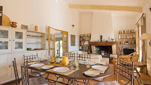 Un coin repas avec une table ronde dressée pour six personnes, entourée de chaises rembourrées. On y trouve une bibliothèque remplie de livres et une cheminée en arrière-plan.