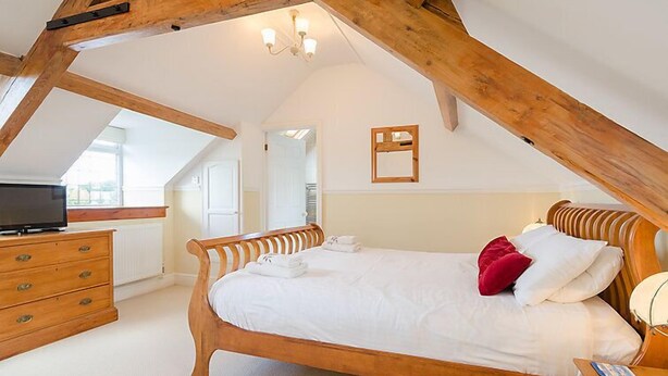 Un dormitorio con techo inclinado, estructura de cama de madera, ropa de cama blanca y un televisor sobre un aparador.