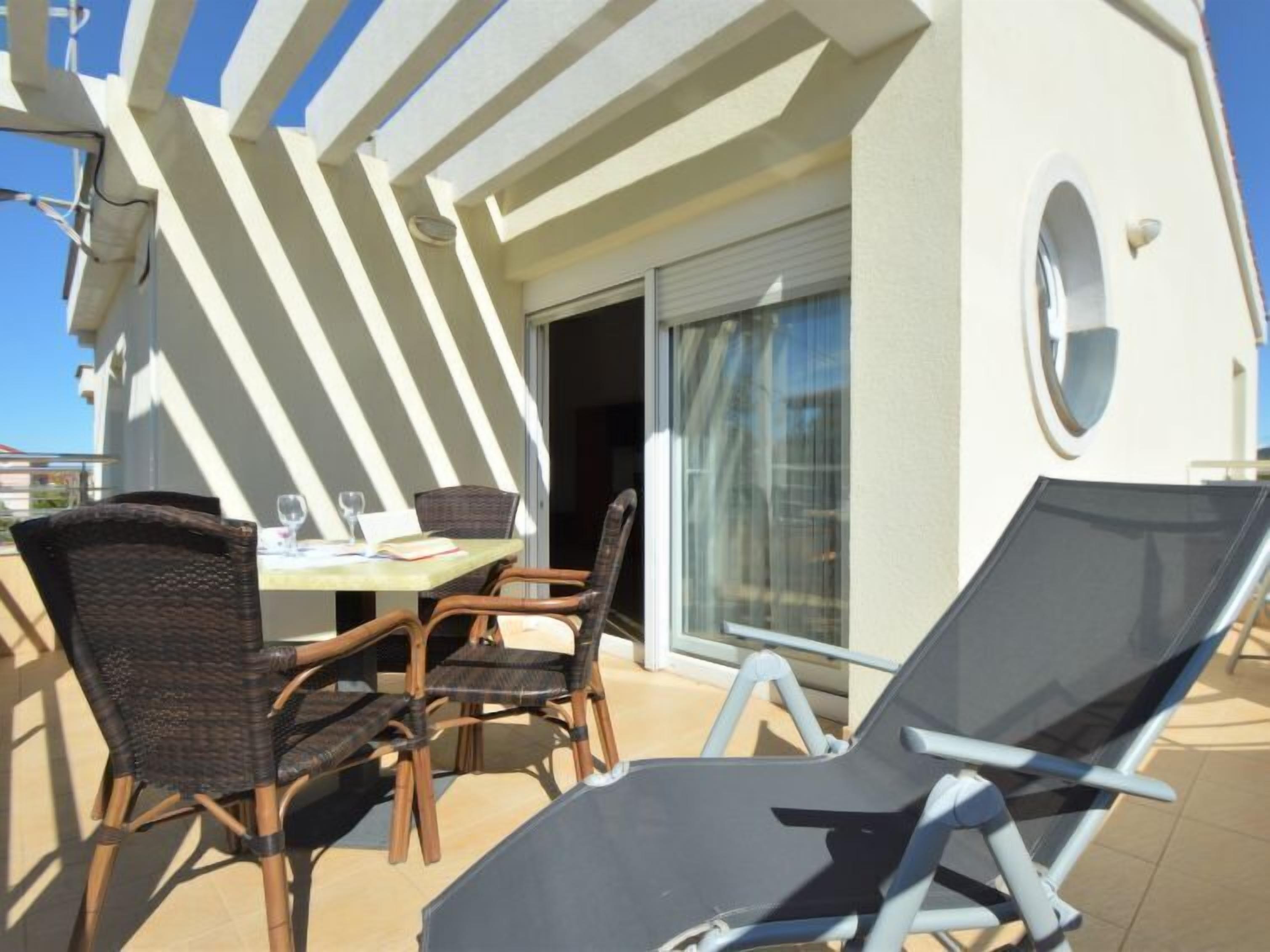 A sunlit outdoor seating area with a table and chairs, a sliding glass door, and a poolside lounge chair.