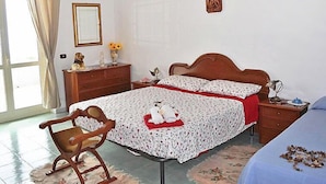 Quarto com uma cama de casal, uma mesinha de cabeceira de madeira, uma cadeira, cômoda e janela com cortinas.