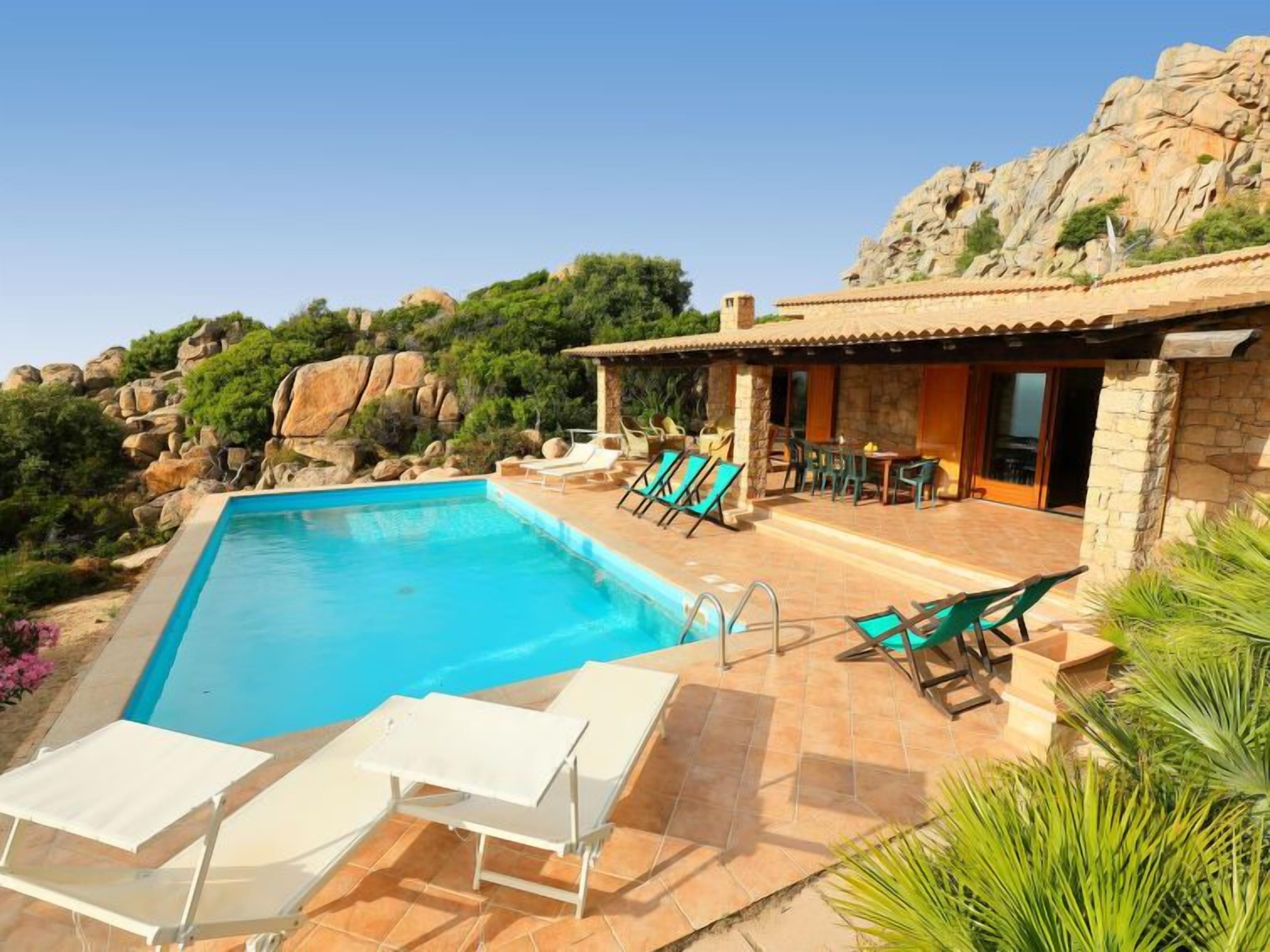 A swimming pool with lounge chairs, a patio area with a table and chairs, and a stone building with a tiled roof.
