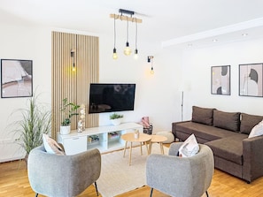 Un soggiorno moderno con una TV a schermo piatto, un divano marrone, due poltrone grigie, una parete con listelli di legno e un tavolino rotondo con un vaso e libri.
