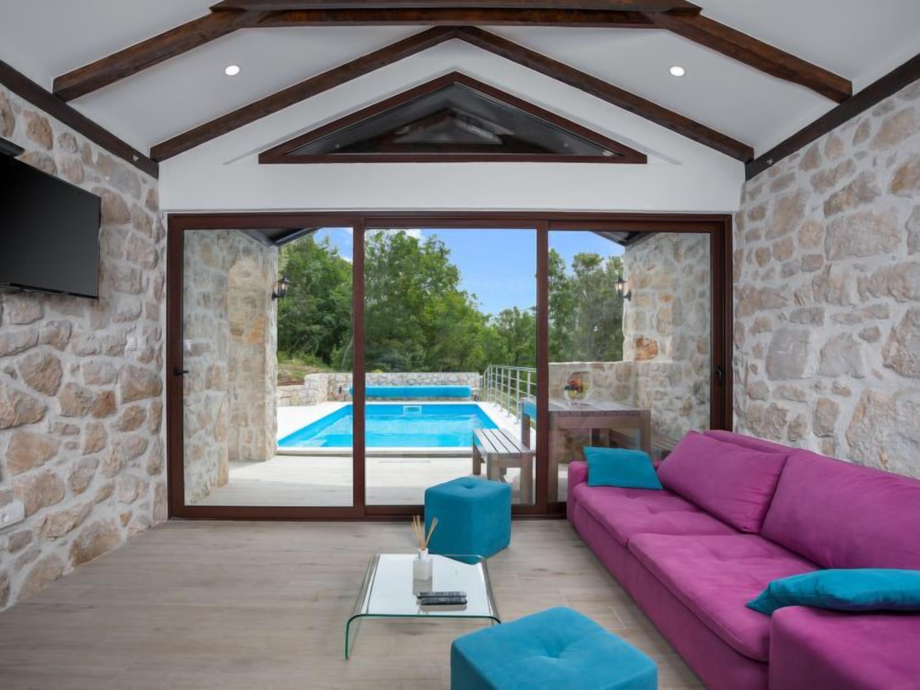 A spacious living area with a stone wall, a large sliding glass door leading to a pool, a pink sofa, and a glass coffee table.