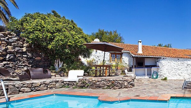 A swimming pool with a stone deck, a barbecue area, and a white building with a tiled roof.