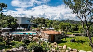 Une maison moderne dotée d’une piscine, de structures en pierre et d’un jardin agrémenté de rochers et d’arbres.
