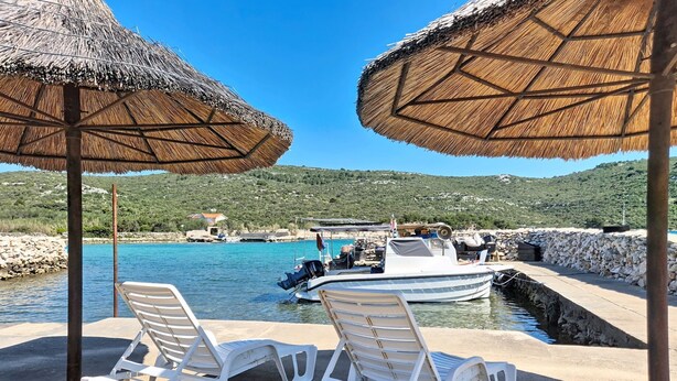 Un cadre idyllique sur la plage, avec deux chaises longues blanches sous des parasols en chaume, un petit bateau amarré près d’un mur de pierre, et une mer bleue et limpide, avec un paysage vallonné en arrière-plan.
