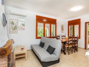 A living room with a sofa, a small table with glasses and a bowl of fruit, a dining area with a wooden table and chairs, and a brick wall.