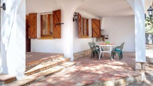A covered patio with a stone floor, a table with chairs, and wooden shutters on the walls. - Villa Pinos by Interhome (L'Ametlla de Mar)