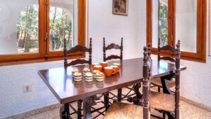 A dining area with a wooden table and chairs, a painting on the wall, and large windows with a view of trees. - Villa Pinos by Interhome (L'Ametlla de Mar)