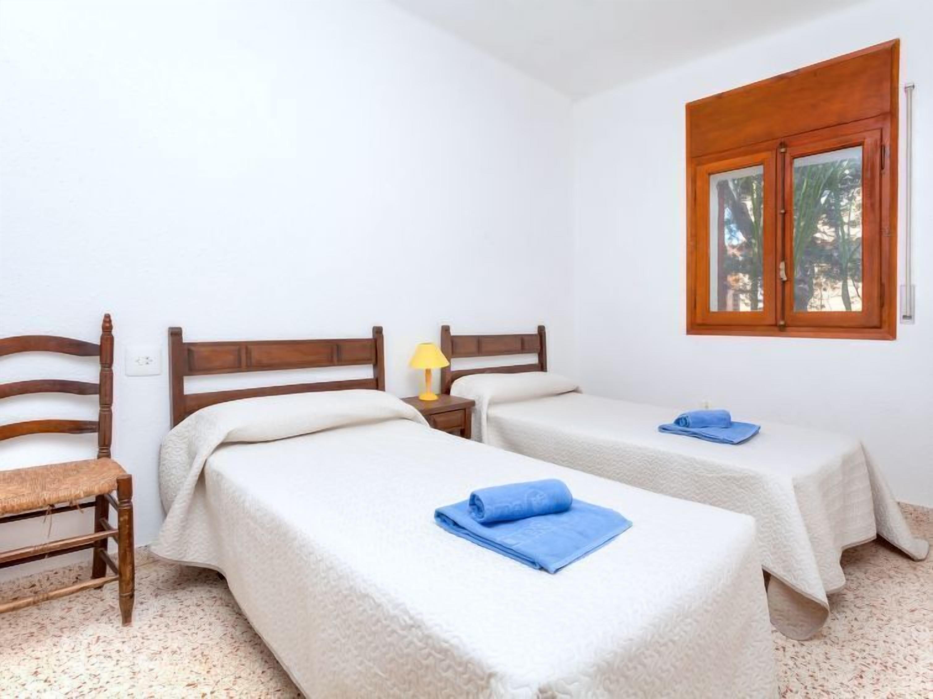 Una habitación con dos camas individuales, una silla de madera, una mesita de noche con una lámpara y una ventana que da a árboles.