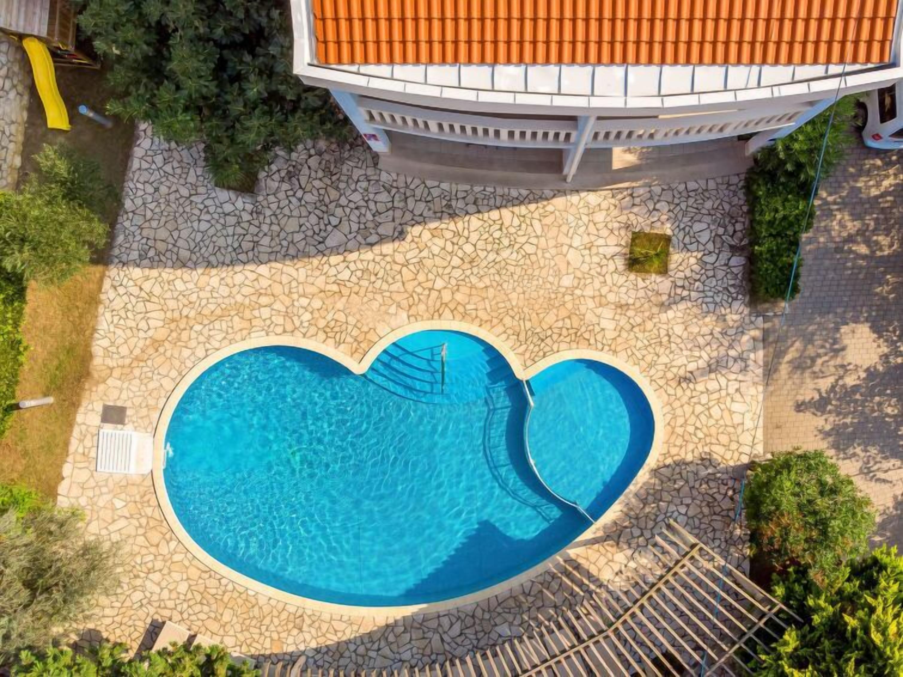 A heart-shaped pool with a stone patio surrounding it, a white building with a tiled roof, and a small garden area with trees and shrubs.