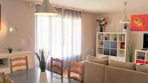 Uma sala de estar moderna com área de jantar, apresentando um sofá, mesa de jantar, cadeiras, luminárias pendentes e uma estante de livros.