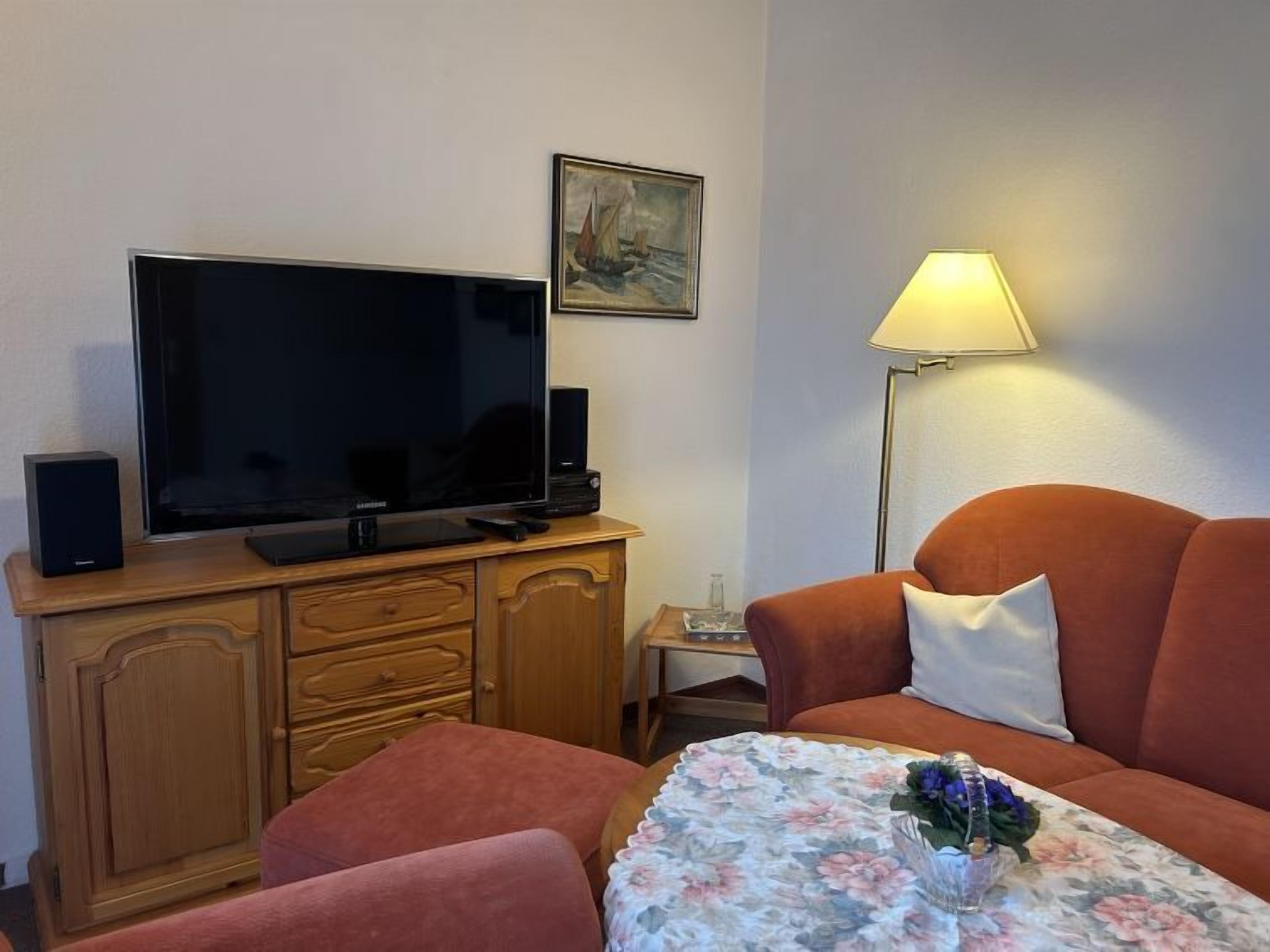 Una sala de estar con televisor de pantalla plana, aparador de madera, sofá naranja y una mesa con mantel floral.
