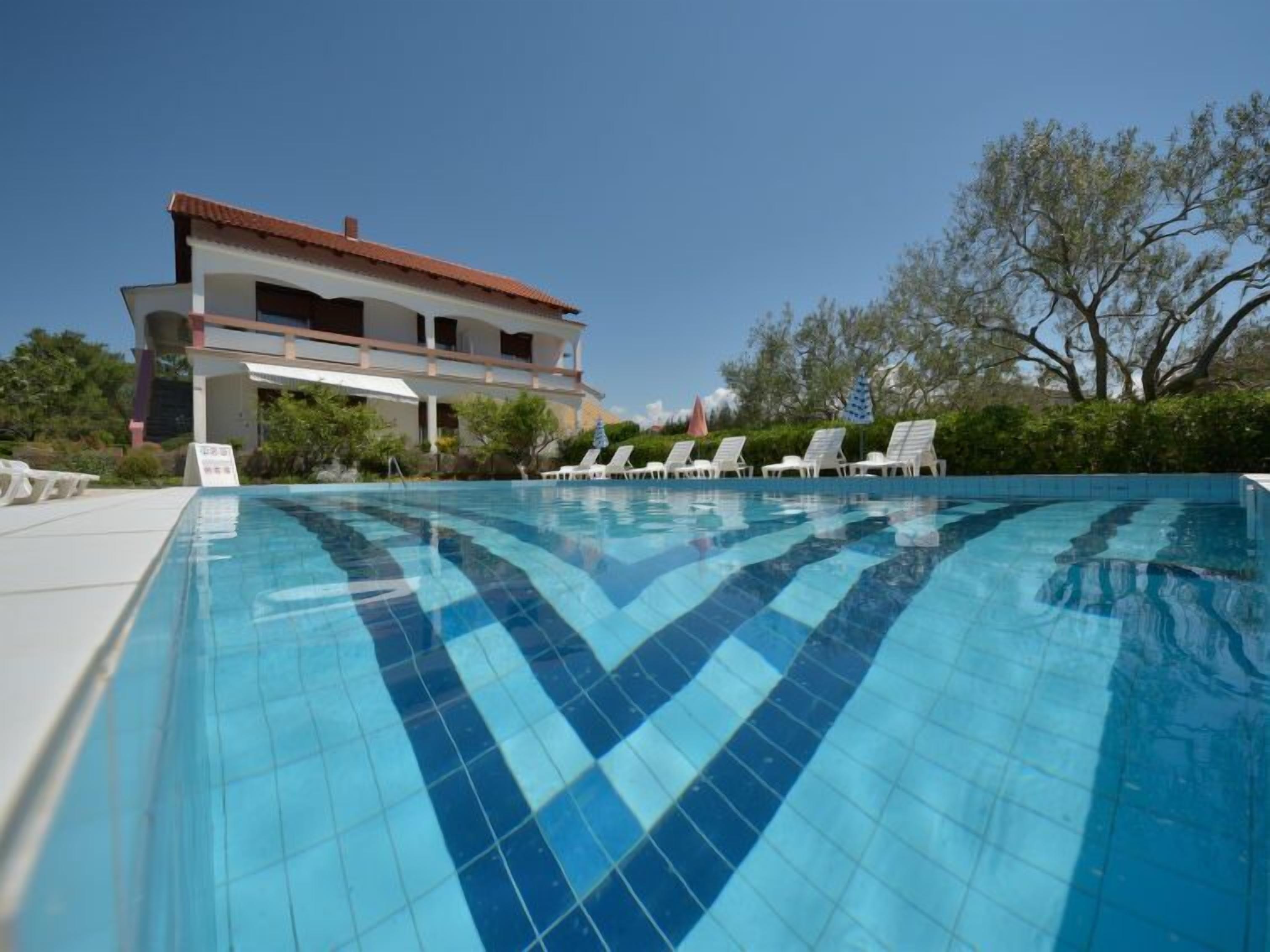 A swimming pool with a tiled pattern, surrounded by lounge chairs and a building with a red roof.