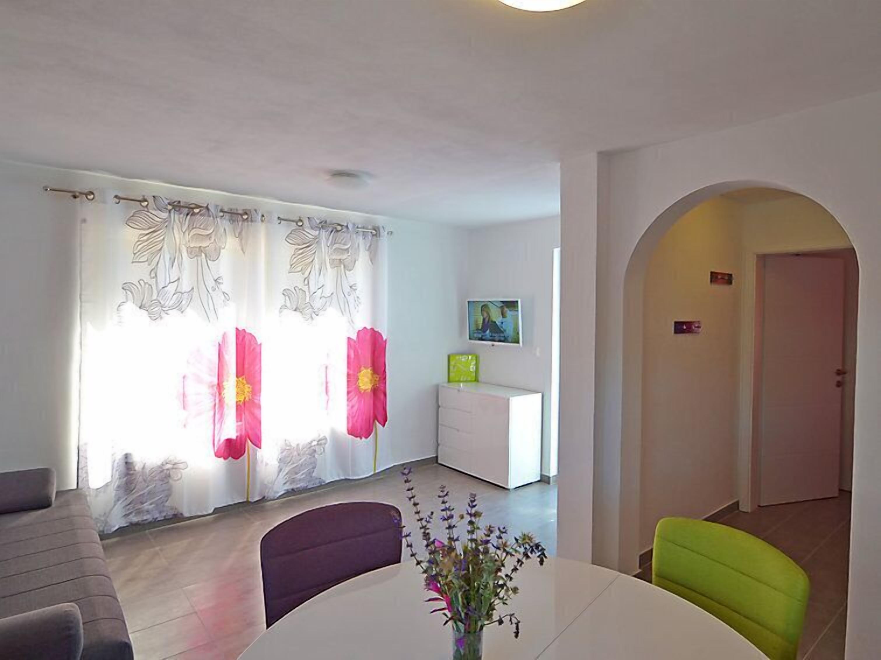 A modern living room with a sofa, a dining table with chairs, a TV on a white cabinet, and a door leading to another room.