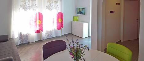 A modern living room with a sofa, a dining table with chairs, a TV on a white cabinet, and a door leading to another room.