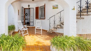 Un patio interior con paredes blancas, suelo de baldosas y dos sillas blancas. Se observa una escalera con barandilla metálica y una puerta con panel de vidrio decorativo.