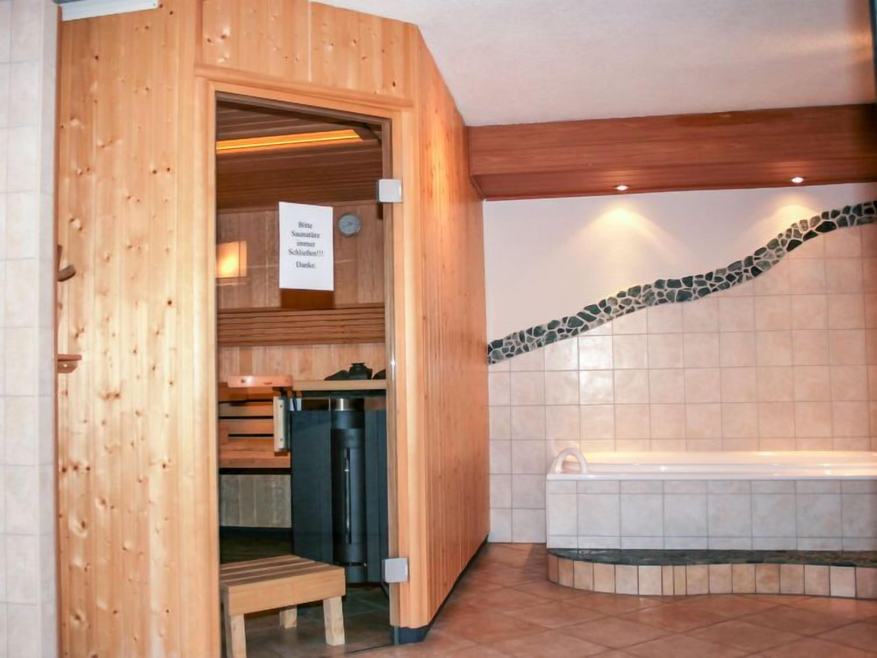 A sauna room with a wooden bench, a bench with a towel, a black heater, and a tiled wall with a mosaic border.
