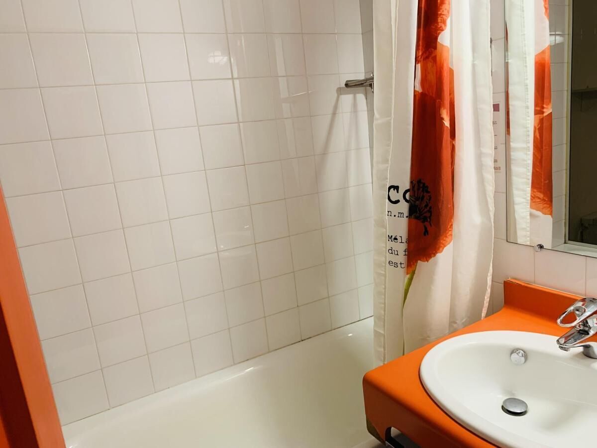 Une salle de bain avec une douche carrelée de carreaux blancs, un lavabo blanc doté d’un plan de toilette orange et un rideau de douche orné de motifs floraux.
