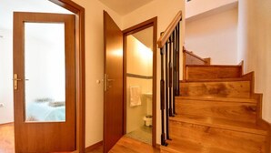 Un escalier en bois menant à l’étage supérieur, une porte en bois avec un panneau en verre dépoli, et une vue sur une salle de bain où une serviette est accrochée au mur.