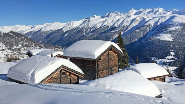 Une cabane en bois, dont le toit est recouvert de neige, entourée de montagnes et d’arbres également couverts de neige.