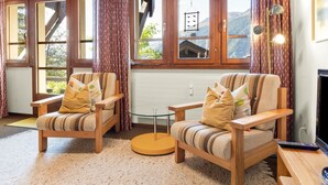 Une pièce confortable avec deux fauteuils en bois, une petite table ronde et un téléviseur. De grandes fenêtres, ornées de rideaux, offrent une vue sur l’extérieur.