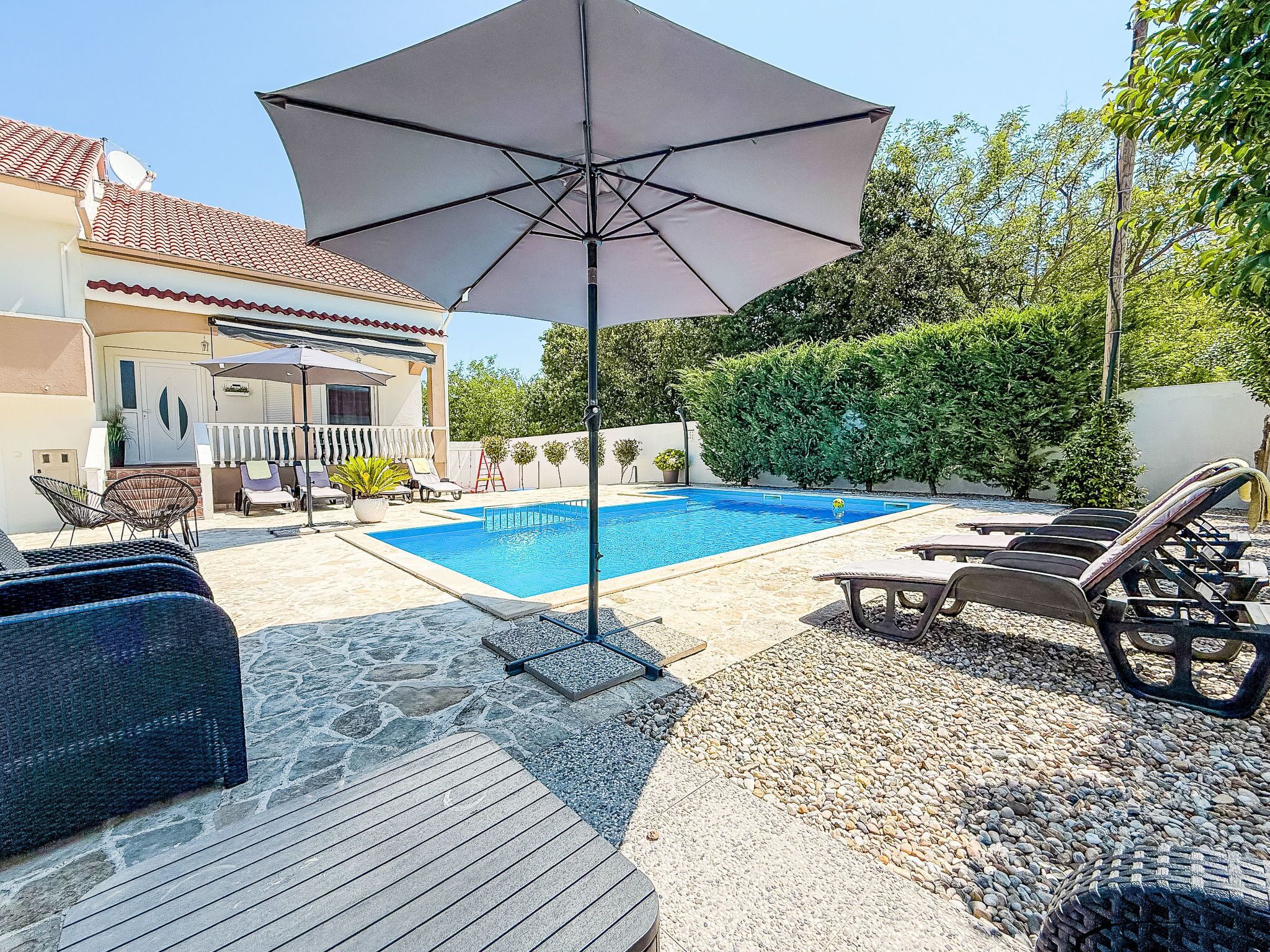 Ein Schwimmbad mit Liegestühlen und Sonnenschirm, umgeben von einer Terrasse mit Steinboden und Blick auf ein Haus mit einem Ziegeldach.