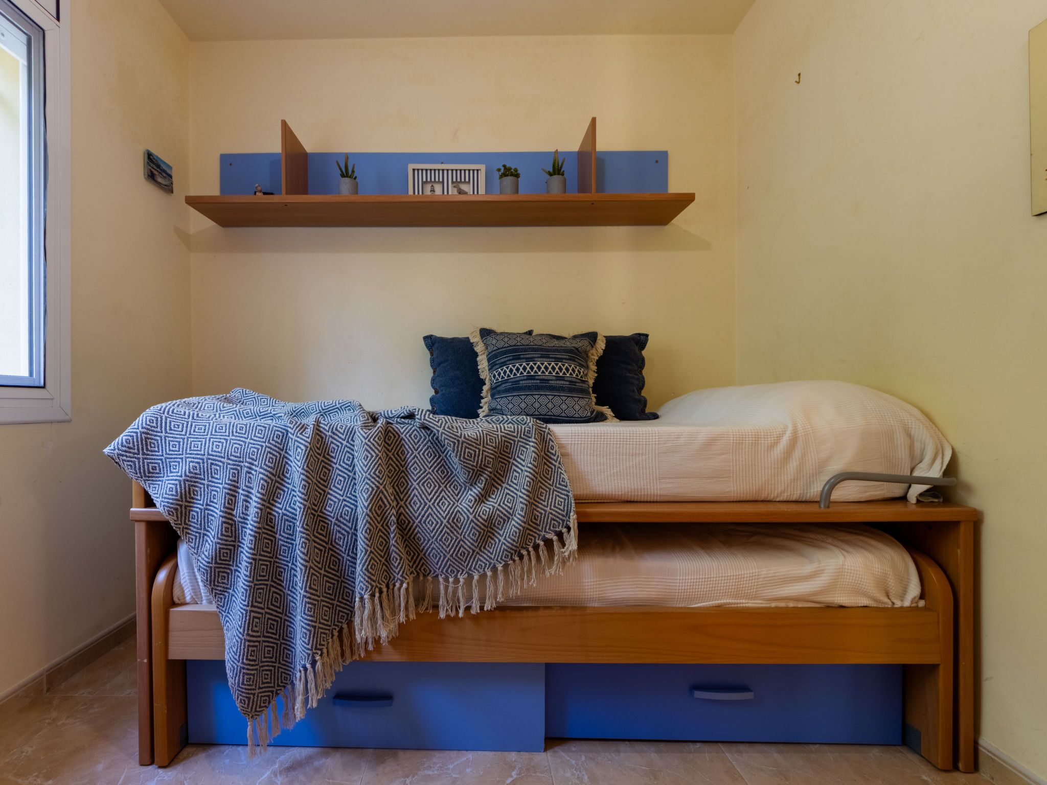 Een knus slaapkamer met een stapelbed, een plank boven het bed en een raam links.