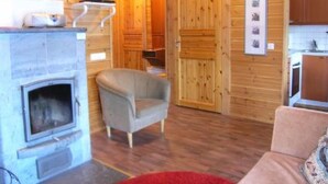 Ein gemütliches Blockhaus-Interieur mit Kamin, einem darüber montierten Fernseher, einem bequemen Sessel und einem durch eine offene Tür einsehbarer Küchenbereich.