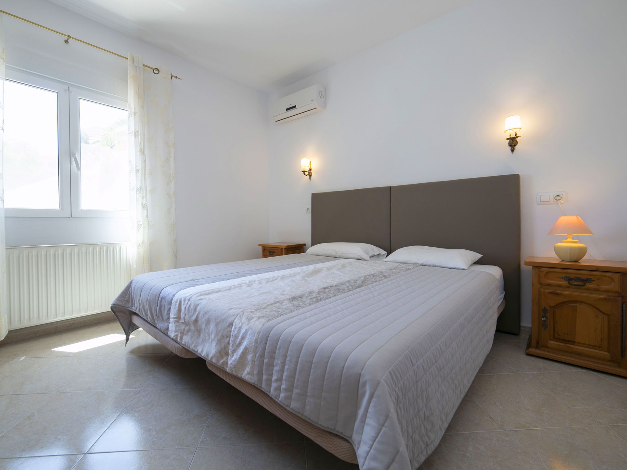 A bedroom with a large bed, two bedside tables with lamps, a window with curtains, and an air conditioning unit on the wall.