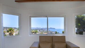 Quarto com sofá bege e duas janelas com vista para uma cidade costeira e oceano.