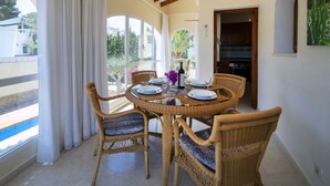 Uma área de refeições com cadeiras de vime e uma mesa posta para quatro, com vista para uma piscina e vegetação.