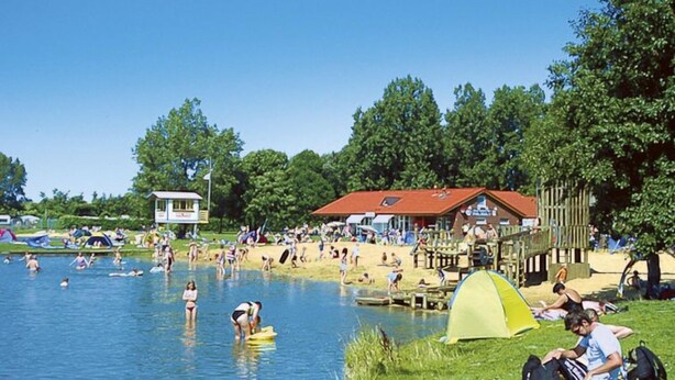 Une scène au bord d’un lac, avec des personnes qui se baignent et prennent des bains de soleil, une tente et un bâtiment avec un toit rouge.