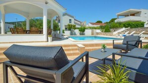 Un espace aménagé au bord de la piscine, avec des sièges d’extérieur, une terrasse couverte et une vue sur la piscine et les bâtiments environnants.