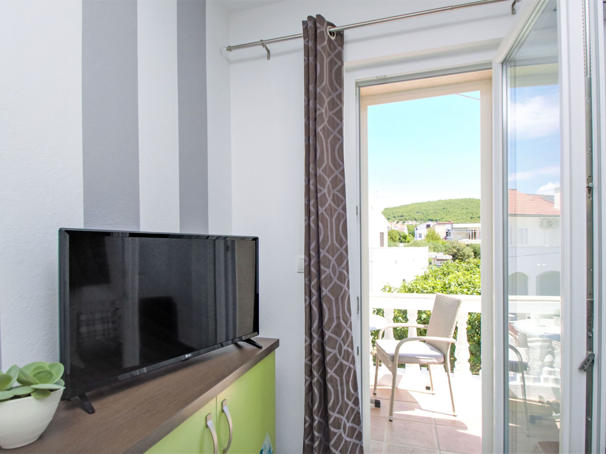 Habitación con televisión en un estante de madera, cortina estampada y vistas a un balcón con una silla y una mesa.
