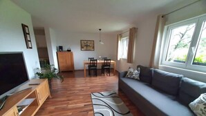 Ett vardagsrum med en soffa, ett tv-appar, ett träskåp, ett matbord, stolar och ett fönster med gardiner.