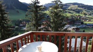 Un balcón con una mesa y una taza, con vistas a un pueblo montañoso con casas y vegetación.