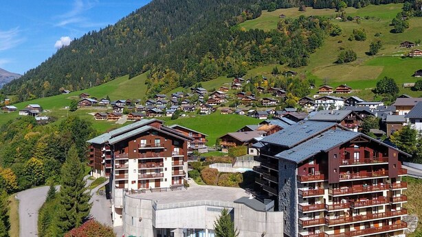 Un pueblo de montaña con edificios de estilo chalete tradicional, una estación de esquí y una vista despejada de las colinas boscosas circundantes.