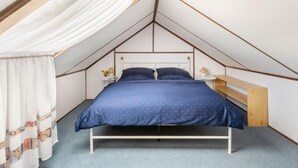 Un dormitorio con techo inclinado, una cama con colcha azul, una mesita de noche con una lámpara y un banco de madera.