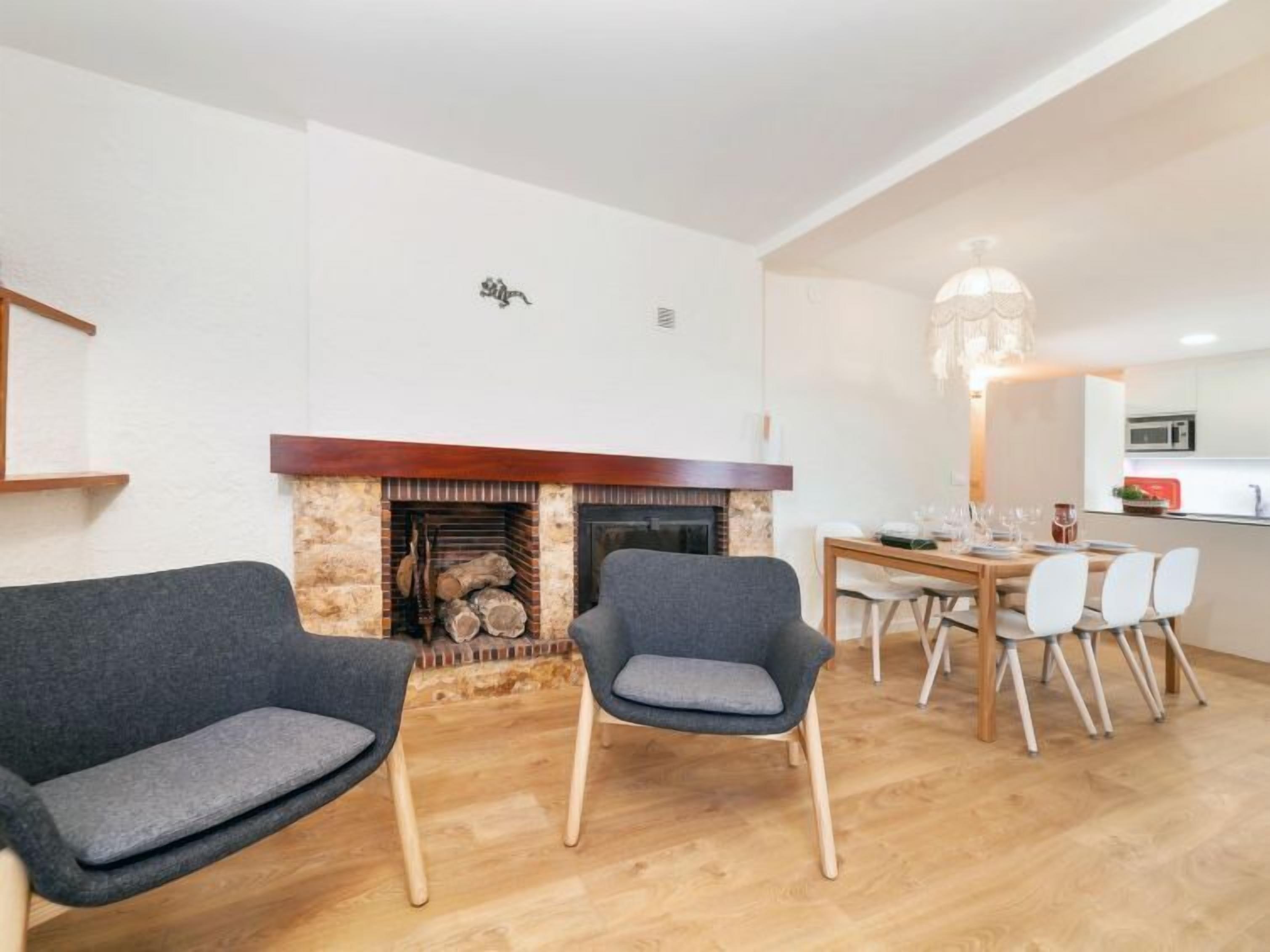 A modern living room with a fireplace, two grey armchairs, a dining table with chairs, and a kitchen in the background.