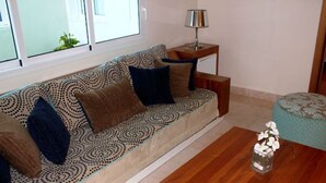 Una sala de estar con un sofá estampado, una mesita auxiliar con una lámpara, una mesa de centro de madera y un jarrón decorativo con flores.