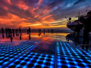 Sonnenuntergang mit Menschen auf einer spiegelnden Fläche, möglicherweise einem Deck oder einer Plattform.