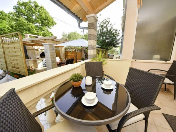 Eine überdachte Terrasse im Freien mit einem runden Glastisch, Korbstühlen und einem kleinen Topfpflanzen.