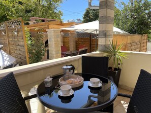 Ein runder Glastisch mit Kaffeekanne, zwei Tassen und einer Schale Gebäck auf einem Balkon mit Korbstühlen, einem Blumentopf und Blick auf eine Terrasse mit Tisch und Stühlen.