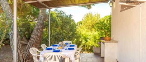 Une terrasse extérieure avec une table dressée pour quatre personnes, entourée de chaises en plastique blanches, sous une pergola en bois.
