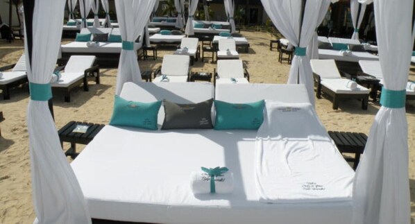 A beachside hotel room with a canopy bed, multiple lounge chairs, and a sandy beach in the background.