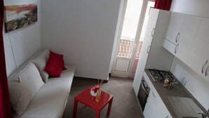 A compact living space with a sofa, a red table with candles, a kitchen with white cabinets, and a balcony door.