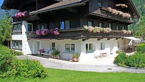 Eine zweistöckige Chalet mit Balkonen, die mit Blumen geschmückt sind, einer überdachten Außenlounge und einem Berg im Hintergrund.