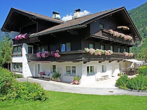Un chalet de dos pisos con balcones decorados con flores, un área de estar al aire libre cubierta y el paisaje de una montaña al fondo.